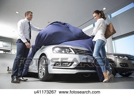 Stock Photo - Salesman uncovering new car to female customer in car dealership. Fotosearch