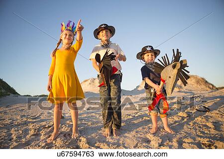 Stock Image - Sister and brothers dressed as native american and cowboys pointing from sand dunes. Fotosearch