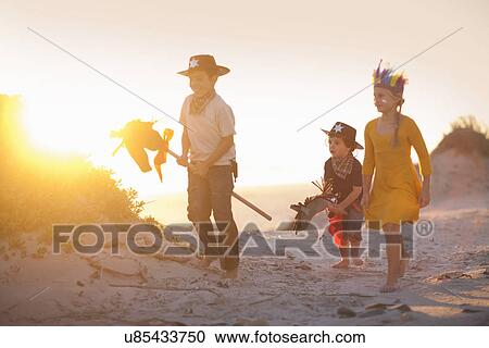 Stock Image - Three children dressed as native american and cowboys in sand dunes. Fotosearch