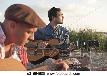 Stock Image - Two adult male friends playing acoustic guitars on Bournemouth beach, Dorset, UK. Fotosearch