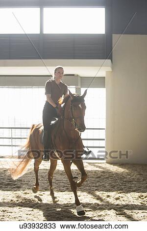Stock Image - Woman trotting on chestnut horse in indoor paddock. Fotosearch