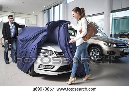 Stock Photo - Woman uncovering new car with boyfriend in car dealership. Fotosearch