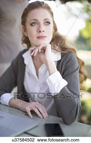 Stock Image - Young businesswoman with hand on chin at garden table. Fotosearch