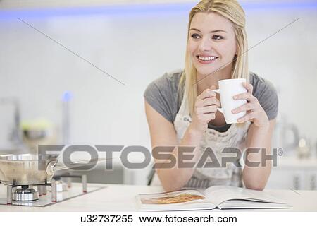 Stock Photography - Young woman drinking coffee and looking at recipe book in kitchen. Fotosearch