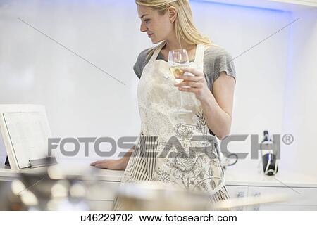 Stock Image - Young woman drinking glass of white wine whilst reading recipe book in kitchen. Fotosearch