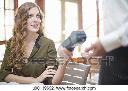 Stock Image - Young woman handing card machine reader to waiter in restaurant. Fotosearch