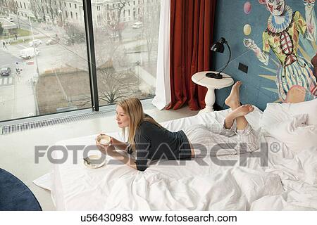 Stock Image - Young woman having a coffee whilst lying on hotel bed. Fotosearch