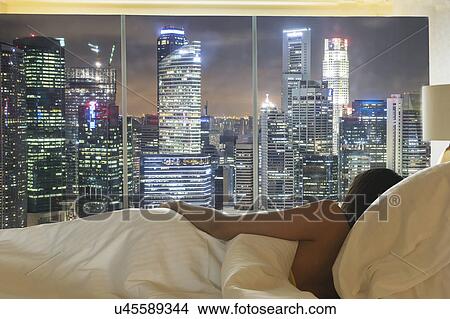 Picture - Young woman lying in bed looking at city skyline through window. Fotosearch