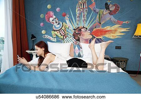 Stock Photograph - Young woman lying on hotel bed texting on smartphone. Fotosearch