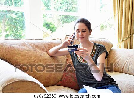 Stock Image - Young woman sitting on sofa home shopping on landline telephone. Fotosearch