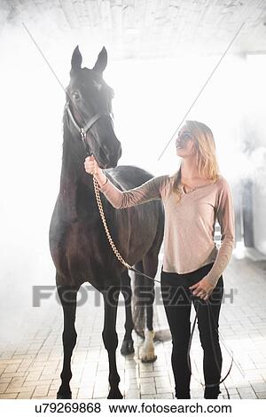 Young woman standing in stables with black horse View Large Photo Image Stock Photo - Young woman standing in stables with black horse. Fotosearch