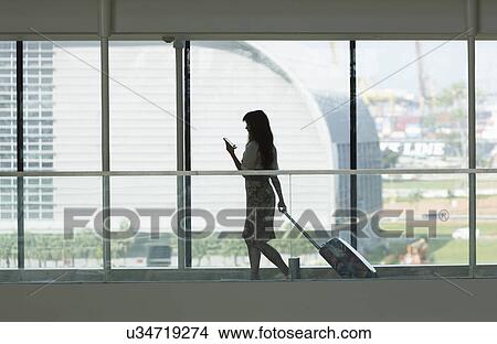 Picture - Young woman using smartphone and pulling suitcase in airport. Fotosearch