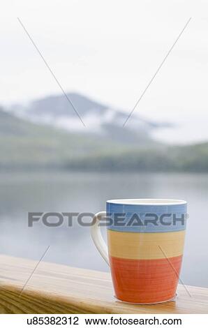 Stock Image - Close-up of mug on balustrade, lake in background. Fotosearch