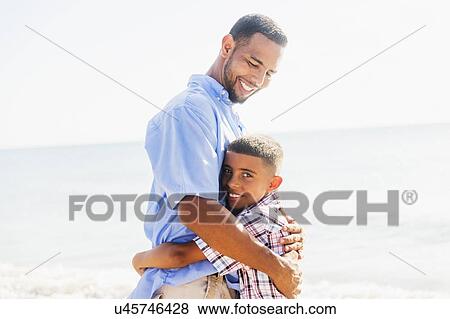 Stock Photo - Father and son (10-11) embracing on beach. Fotosearch