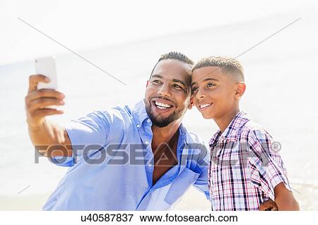 Stock Photo - Father and son (10-11) photographing self on beach. Fotosearch