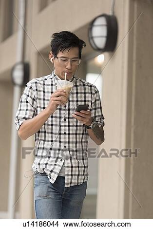 Picture - Man walking on sidewalk with iced coffee and mobile phone. Fotosearch