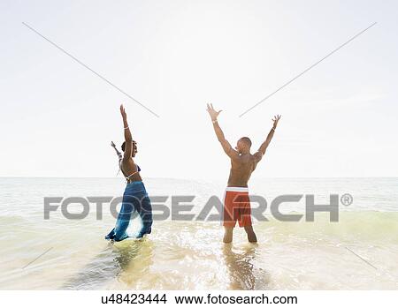 Picture - Mature couple standing in sea with arms raised. Fotosearch