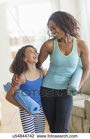 Stock Image - Mother and daughter (8-9) holding yoga mats in living room. Fotosearch