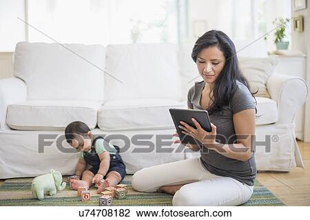 Stock Image - Mother using tablet pc while her son (6-11 months) playing with building blocks. Fotosearch