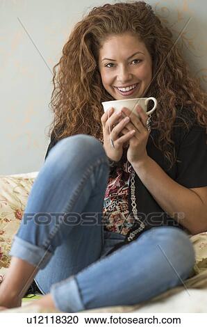 Stock Image - Portrait of woman sitting on bed with tea cup. Fotosearch