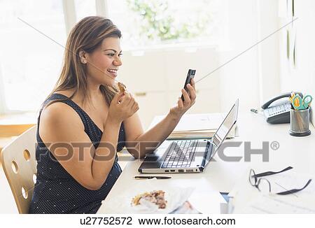 Stock Image - Woman eating croissant and using mobile phone in office. Fotosearch
