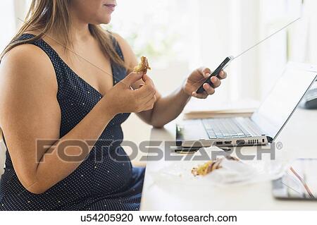 Stock Image - Woman eating croissant and using mobile phone in office. Fotosearch