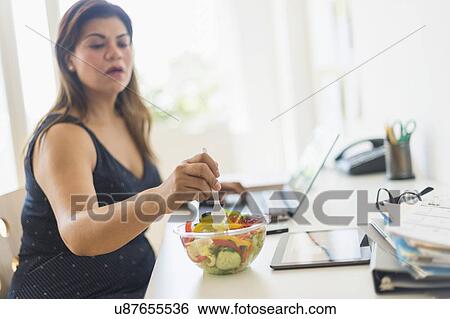 Stock Photograph - Woman eating salad and using laptop in office. Fotosearch