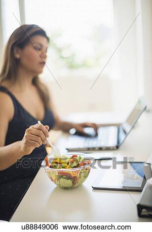 Stock Photograph - Woman eating salad and using laptop in office. Fotosearch