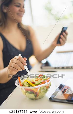 Stock Image - Woman eating salad and using mobile phone in office. Fotosearch