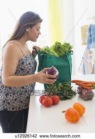Stock Image - Woman taking out vegetables of grocery bag. Fotosearch