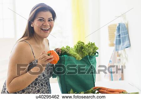 Stock Image - Woman taking out vegetables of grocery bag. Fotosearch
