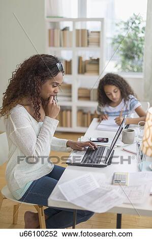 Stock Image - Woman using laptop at home, girl (8-9) doing homework in background. Fotosearch
