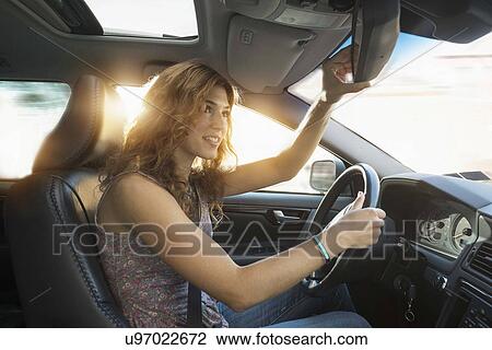 Stock Image - Young woman adjusting rear view mirror while driving. Fotosearch