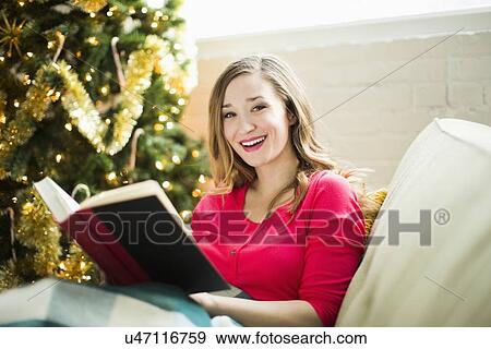 Stock Photo - Young woman reading book on sofa at Christmas time. Fotosearch