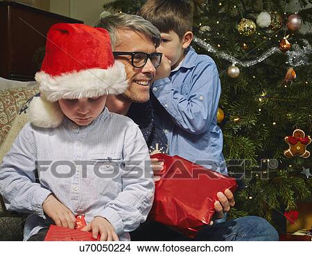 Boy whispering to father whilst brother opens christmas gift View Large Photo Image Picture - Boy whispering to father whilst brother opens christmas gift. Fotosearch