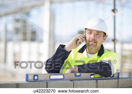 Stock Image - Builder with spirit level on construction site chatting on smartphone. Fotosearch