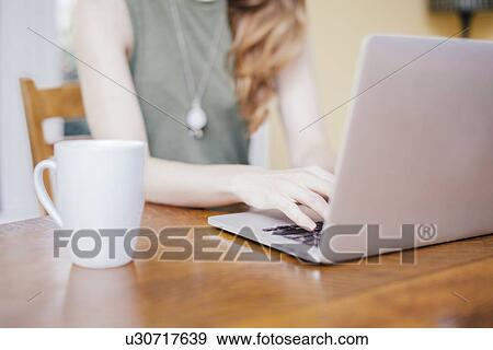 Stock Photo - Cropped shot of young woman typing on laptop at dining room table. Fotosearch