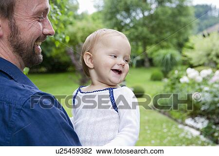 Stock Image - Father carrying baby daughter in garden. Fotosearch