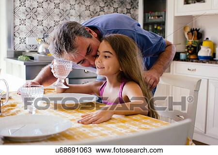 Stock Image - Father kissing daughter on cheek at kitchen table. Fotosearch