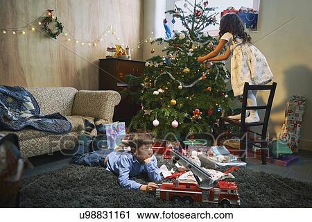 Girl arranging christmas tree whilst brother plays with xmas gifts on sitting room floor View Large Photo Image Stock Image - Girl arranging christmas tree whilst brother plays with xmas gifts on sitting room floor. Fotosearch