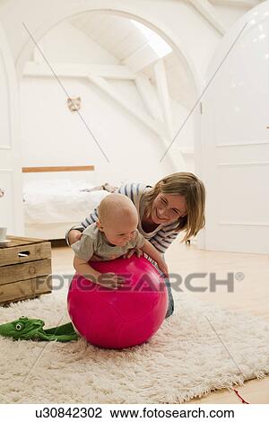 Stock Image - Mature mother and baby daughter on top of exercise ball in sitting room. Fotosearch