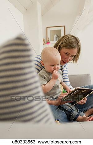 Stock Image - Mature mother and baby daughter reading storybook on sitting room sofa. Fotosearch
