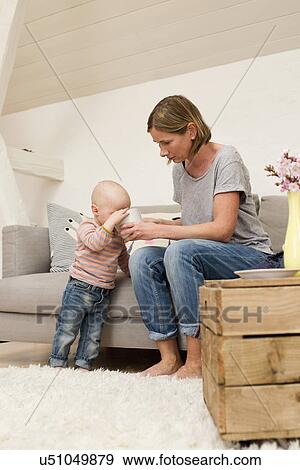 Stock Photo - Mature mother sharing drink with baby daughter in sitting room. Fotosearch