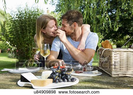 Stock Photo - Mid adult man feeding girlfriend from picnic table in garden. Fotosearch