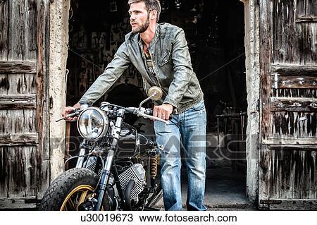 Stock Image - Mid adult man pushing motorcycle out of barn. Fotosearch
