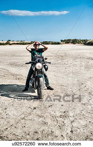 Stock Image - Mid adult man sitting on motorcycle on arid plain, Cagliari, Sardinia, Italy. Fotosearch