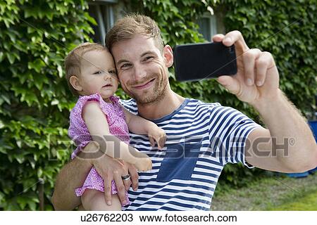Stock Image - Mid adult man with baby daughter taking selfie on smartphone in garden. Fotosearch