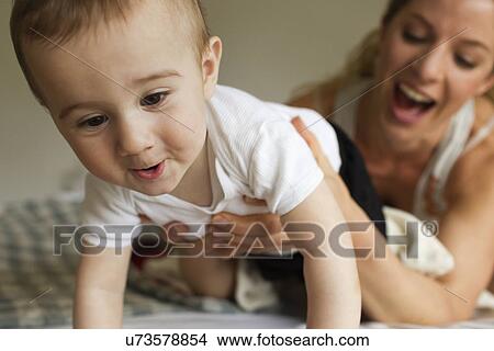 Picture - Mother guiding baby boy crawling on bed. Fotosearch