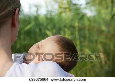 Over shoulder portrait of mid adult mother and sleeping baby daughter in garden View Large Photo Image Picture - Over shoulder portrait of mid adult mother and sleeping baby daughter in garden. Fotosearch