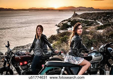 Stock Image - Portrait of two female motorcyclist friends on coast at sunset, Cagliari, Sardinia, Italy. Fotosearch